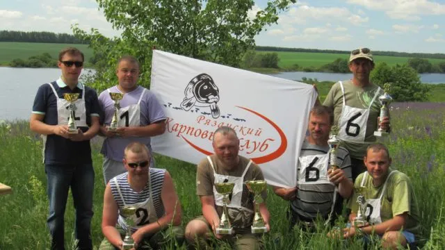 Победители соревнований по карпфишингу с кубками на фоне флага карпового клуба
