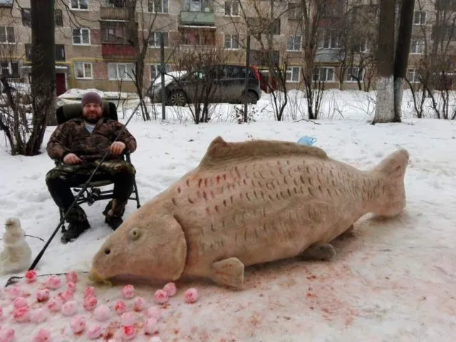 Рыбак со снежной скульптурой карпа во дворе жилого дома зимой
