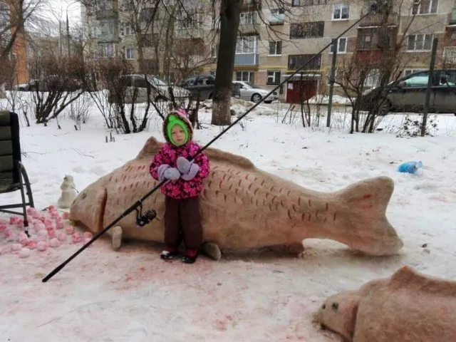 Ребенок с удочкой рядом со снежной скульптурой карпа во дворе жилого дома